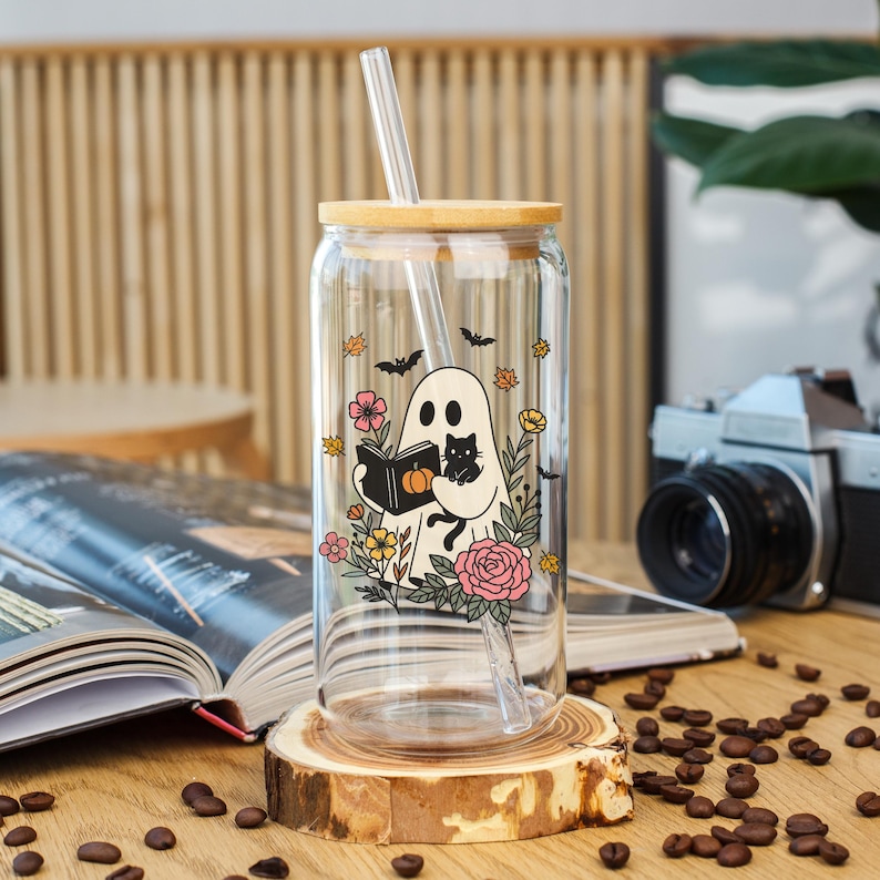 May include: A clear glass can-shaped tumbler with a bamboo lid and straw. The tumbler features a whimsical ghost design holding a book and a black cat, surrounded by flowers and bats. The tumbler is displayed on a wooden coaster.