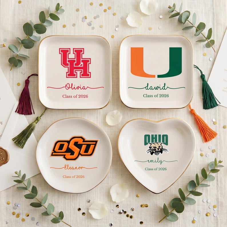 Puede incluir: Cuatro platos de cer&aacute;mica personalizados con bordes dorados. Cada plato presenta un logotipo universitario, un nombre y "Class of 2026". Las formas incluyen un cuadrado, un redondo y un coraz&oacute;n. Borlas decorativas y vegetaci&oacute;n rodean los platos.