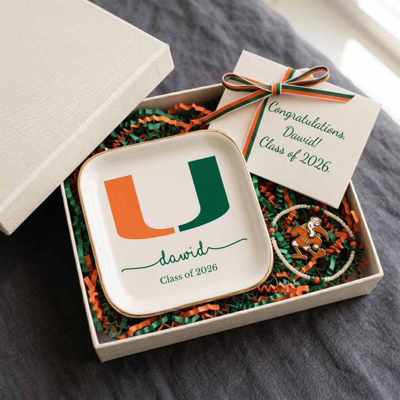 Puede incluir: Caja de regalo abierta con tapa blanca, que contiene una bandeja de baratijas blanca con ribete dorado, una pulsera y una tarjeta. La bandeja tiene el texto "david" y "Class of 2026" debajo de un logotipo "U" verde y naranja. La tarjeta dice "&iexcl;Felicidades, David! Class of 2026."
