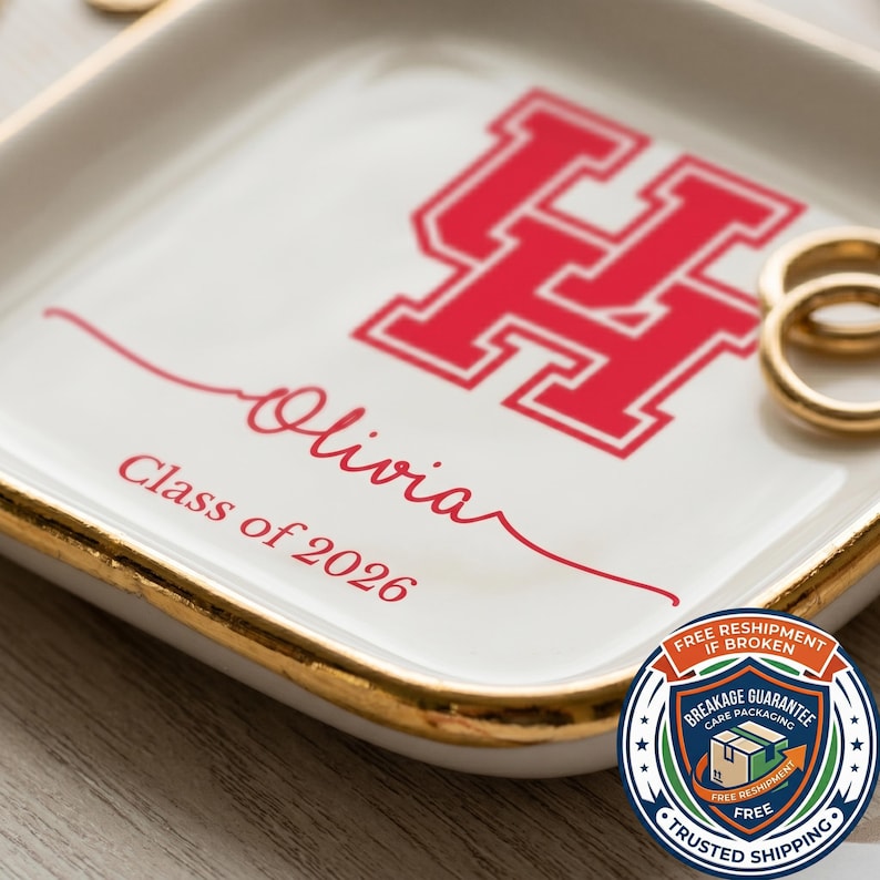 Puede incluir: Una bandeja de cer&aacute;mica blanca con borde dorado, con un logotipo "UH" rojo y el nombre "Olivia" con "Class of 2026" en escritura roja. La bandeja est&aacute; dise&ntilde;ada para guardar objetos peque&ntilde;os como anillos.