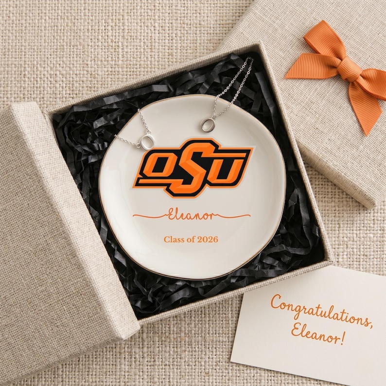 Puede incluir: Un plato de cer&aacute;mica blanco con el logotipo de la Universidad Estatal de Oklahoma en naranja y negro, personalizado con un nombre y el a&ntilde;o de graduaci&oacute;n. El plato est&aacute; en una caja de regalo con un lazo a juego y una tarjeta que dice "&iexcl;Felicidades, Eleanor!" Tambi&eacute;n se incluyen dos collares de plata.