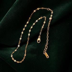 Può includere: Una delicata collana color oro con una piccola gemma rotonda e trasparente. La collana presenta una catena di piccole maglie piatte di forma ovale e una catena separata con chiusura a moschettone. Il gioiello è esposto su una superficie di velluto verde scuro.