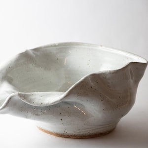 May include: A handmade ceramic bowl with a unique, ruffled edge. The bowl is white with subtle speckles and a natural, unglazed base. The design is organic and artistic, suitable for serving or display.