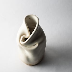 May include: A white ceramic vase with an irregular, pinched, and folded rim design. The body of the vessel features a smooth, matte surface with soft, organic curves and indentations that create a unique, asymmetrical silhouette from the top view.