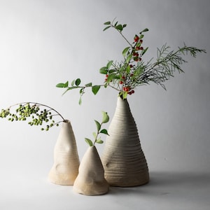 May include: Three off-white ceramic vases of varying sizes, each holding different types of greenery. The largest vase has a textured, ridged design and holds a branch with red berries. The other two vases have a smooth, organic shape.