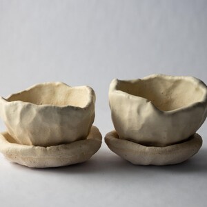 May include: Two off-white ceramic bowls with textured surfaces, each sitting on a matching saucer. The bowls have irregular, organic shapes, suggesting a handmade quality. The neutral color palette and simple design create a minimalist aesthetic.
