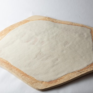 May include: A handmade, irregularly shaped serving board with a light beige, textured surface. The board has a raised, decorative border with a floral pattern in a darker beige color. The center is a lighter, smooth surface.