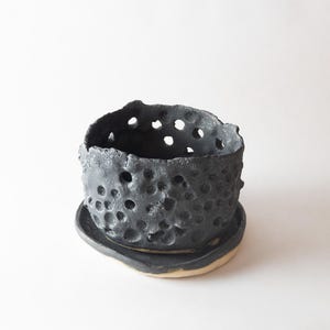 May include: A dark gray ceramic planter with a textured surface and numerous holes. The planter sits on a matching saucer with a light beige base. The handmade pot has a unique, organic shape, suitable for small plants.