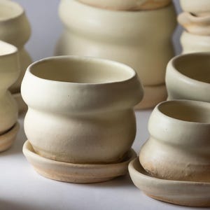 May include: A collection of off-white ceramic planters with a rounded, bulbous shape and a smooth matte finish. Each planter sits on a matching circular saucer, featuring a wide rim and a slightly tapered base, arranged in a group.