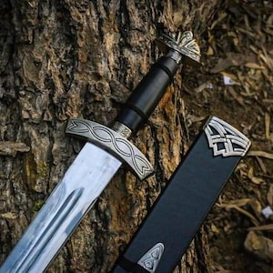 Op de afbeelding: Een Vikingzwaard met een zilveren lemmet en een sierlijk handvat. Het zwaard heeft een zwarte leren grip en wordt getoond met zijn zwarte schede, die zilveren details heeft. Het zwaard ligt op een getextureerde boomstam.