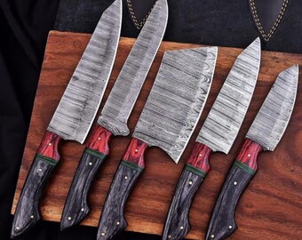 Puede incluir: Un juego de seis cuchillos de cocina de acero de Damasco con mangos de madera. Los mangos son rojos, negros y verdes. Las hojas tienen un patrón único. Los cuchillos están colocados sobre una tabla de cortar de madera.