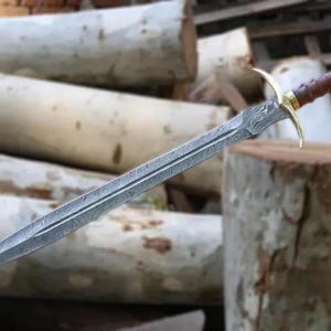 Könnte beinhalten: Ein detailliertes, silberfarbenes Schwert mit gemusterter Klinge und einem braunen, lederumwickelten Griff. Der Knauf hat goldfarbene Akzente. Das Schwert wird vor dem Hintergrund von Holzstämmen präsentiert, was ein mittelalterliches oder Fantasy-Thema suggeriert.