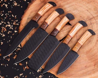 May include: A set of five kitchen knives with black blades and wooden handles. The blades have a textured, hammered appearance. The handles are a light wood color with dark wood accents and black bolsters. The knives are arranged on a wooden cutting board.