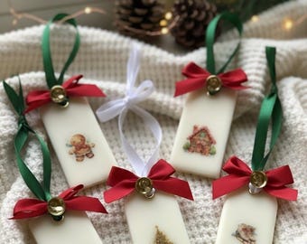 May include: Cream-coloured, rectangular Christmas ornaments, each with a festive illustration. They feature a red bow, green ribbon, and a small gold bell. Designs include a gingerbread man, a house, a Christmas tree, a snowman, and ice skates.
