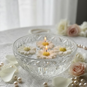 Puede incluir: Un cuenco de vidrio transparente lleno de agua y velas flotantes con forma de margarita. Dos velas están encendidas, con centros amarillos y pétalos blancos. El cuenco está sobre un mantel de encaje blanco, con perlas y pétalos de rosa esparcidos.