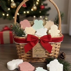 Puede incluir: Una cesta de mimbre llena de jabones con temática navideña en varias formas y colores, incluyendo un muñeco de nieve, un árbol de Navidad y un hombre de jengibre. La cesta está adornada con un lazo rojo y una etiqueta que dice "Season's Greetings".