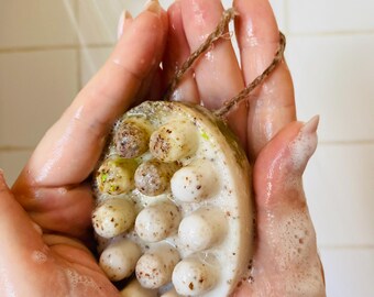 May include: Oval-shaped soap with textured, rounded protrusions and a natural rope loop. The soap is held in wet hands, with visible suds and water droplets. The soap has a light beige color with darker speckles.
