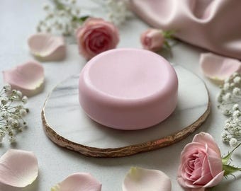 以下が含まれることがあります： 丸い薄いピンク色の石鹸が、ピンクのバラの花びら、小さな白い花、ピンクのバラに囲まれた、大理石と金の縁取りのトレイの上に置かれています。背景にはピンクのサテン生地があります。