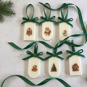 以下が含まれることがあります： 緑のリボンが付いた白いクリスマスオーナメントが6つ。それぞれに異なるお祝いの画像が描かれています。くるみ割り人形、サンタクロース、雪だるま、テディベア、クリスマスツリー、ジンジャーブレッドハウス。白い表面に配置されています。