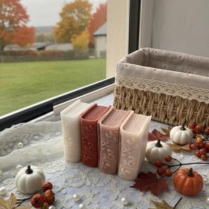 Puede incluir: Cuatro velas con forma de libro en tonos crema, rojo y rosa, dispuestas sobre un mantel de encaje blanco. La escena incluye pequeñas calabazas, bayas y hojas de otoño, con una cesta de mimbre en el fondo.