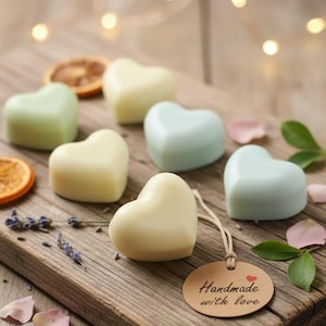 May include: Heart-shaped soaps in pastel colors, including green, blue, and cream, arranged on a wooden board. A small tag reads "Handmade with love." Dried orange slices, lavender sprigs, and rose petals add to the aesthetic. The soaps are a charming gift idea.