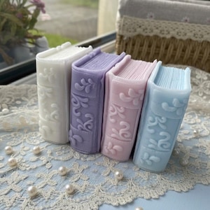 Könnte beinhalten: Vier seifenförmige Bücher in Weiß, Lila, Rosa und Hellblau. Jede Seife hat ein geprägtes Blumenmuster. Sie sind auf einer hellblauen Spitzendecke mit verstreuten Perlen angeordnet.