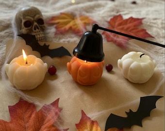 Könnte beinhalten: Ein Halloween-Stillleben mit einem Totenkopf, kürbisförmigen Kerzen und Herbstblättern. Im Mittelpunkt steht eine orangefarbene Kürbiskerze mit einer schwarzen Laterne, umgeben von weißen und orangefarbenen Kerzen, Fledermäusen und dekorativen Elementen.