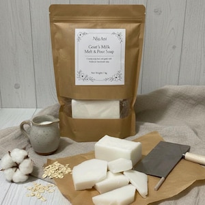 May include: A brown paper bag labelled "NiaAni Goat's Milk Melt & Pour Soap" with a clear window, containing white soap. Several cut pieces of white soap, a small jug, oats, a cotton ball, and a cleaver are also present.