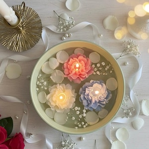 Peut inclure: Une composition décorative avec un bol vert clair rempli d'eau et trois bougies en forme de fleurs allumées, de couleurs rose, jaune et bleu. Des pétales blancs et du gypsophile entourent les bougies, avec un bougeoir doré et un ruban blanc en arrière-plan.