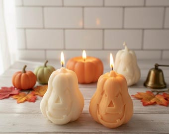 May include: Two lit pumpkin-shaped candles with carved faces, one cream and one orange, sit on a wooden surface. Other pumpkin-shaped candles in orange and green are in the background, along with fall leaves. A small brass bell is also visible.