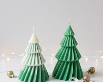 May include: Two Christmas tree-shaped candles. One is green and white, the other is solid green. Both have a tiered design and a wick at the top. Gold ornaments and a white ribbon with a silver bow are placed around the candles. The background is white.