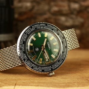 Könnte beinhalten: Eine Vintage-Armbanduhr mit grünem Zifferblatt und goldenen Zeigern, mit einem Metallarmband. Das Zifferblatt zeigt Datum und Uhrzeit, umgeben von einer Lünette mit Städtenamen. Die Uhr liegt auf einer Holzoberfläche.