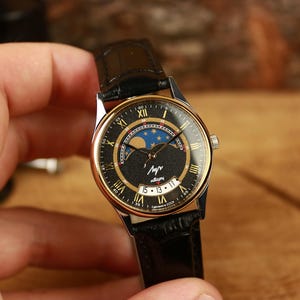 Könnte beinhalten: Vintage Armbanduhr mit schwarzem Zifferblatt, goldfarbener Lünette und schwarzem Lederarmband. Das Zifferblatt der Uhr zeigt römische Ziffern, eine Mondphasenanzeige und kyrillischen Text. Die Uhr wird von einer Person gehalten.