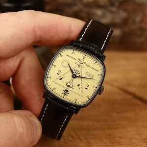 Puede incluir: Un reloj de pulsera vintage con esfera cuadrada y correa de cuero negro. La esfera del reloj es de color crema con marcas negras, incluidas las palabras "МИРОВАЯ КОЛЛЕКЦИЯ" y varios símbolos. El reloj está sujeto por una mano.