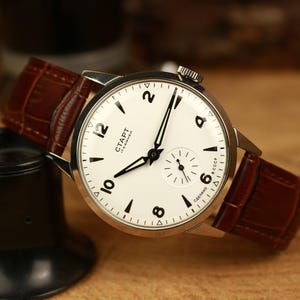 May include: A classic wristwatch featuring a white dial with black numerals and hands, and a small seconds dial. It has a brown leather strap. The watch face displays "CTAPT" and "CCCP". The watch is a timeless accessory.