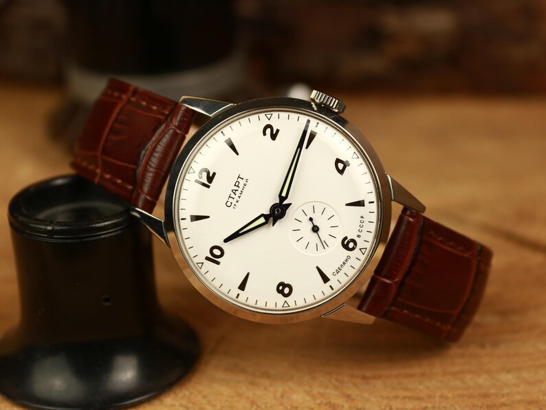 May include: A classic wristwatch featuring a white dial with black numerals and hands, and a small seconds dial. It has a brown leather strap. The watch face displays "CTAPT" and "CCCP". The watch is a timeless accessory.
