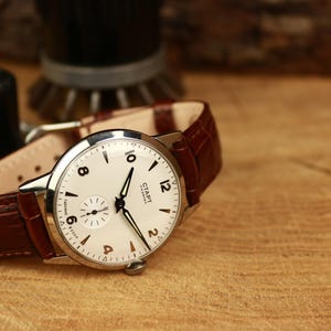 May include: A vintage wristwatch with a white face, black hands, and Arabic numerals. The watch has a small seconds dial and a brown leather strap. The watch is set on a wooden surface.