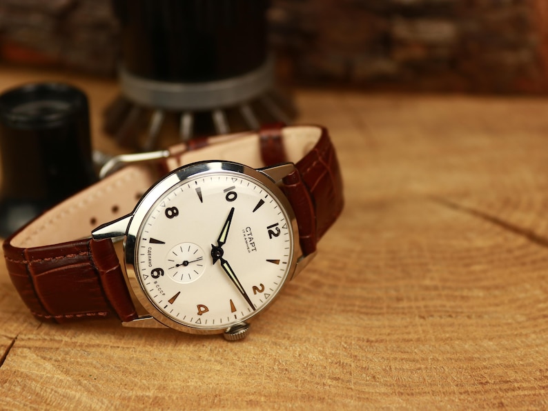 May include: A vintage wristwatch with a white face, black hands, and Arabic numerals. The watch has a small seconds dial and a brown leather strap. The watch is set on a wooden surface.