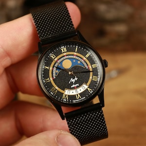 Puede incluir: Reloj de pulsera negro con una correa de malla. La esfera del reloj presenta un diseño de fase lunar con detalles dorados y azules, números romanos y la fecha. La caja y la correa son negras.