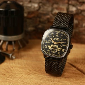 May include: A black wristwatch with a square face and a mesh strap. The watch face is black with gold numerals and symbols. The watch is set against a wooden background, with watchmaking tools in the background.