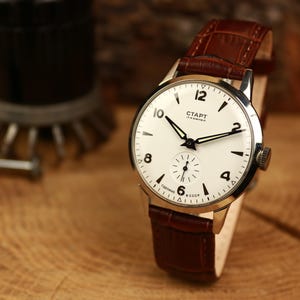 May include: A vintage wristwatch with a white face, black numerals, and black hands. The watch has a brown leather strap and a silver-coloured case. The watch face has the word "CTAPT" printed on it. The watch is displayed on a wooden surface.