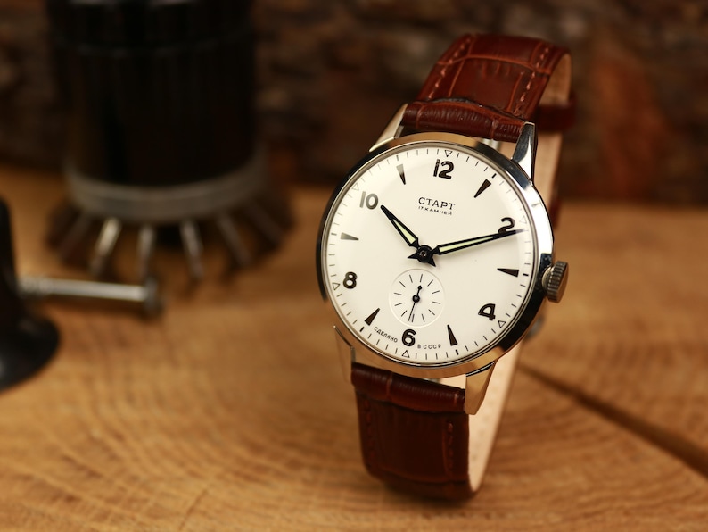 May include: A vintage wristwatch with a white face, black numerals, and black hands. The watch has a brown leather strap and a silver-coloured case. The watch face has the word "CTAPT" printed on it. The watch is displayed on a wooden surface.