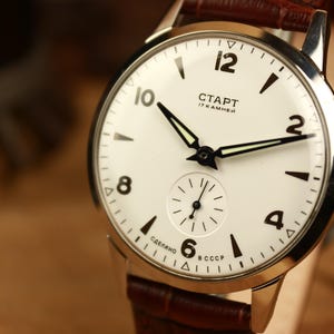 May include: A vintage wristwatch with a white face, black numerals, and black hands. The watch features a small seconds dial and a brown leather strap. The watch face displays the Cyrillic text "CTAPT" and "17 KAMHEЙ".