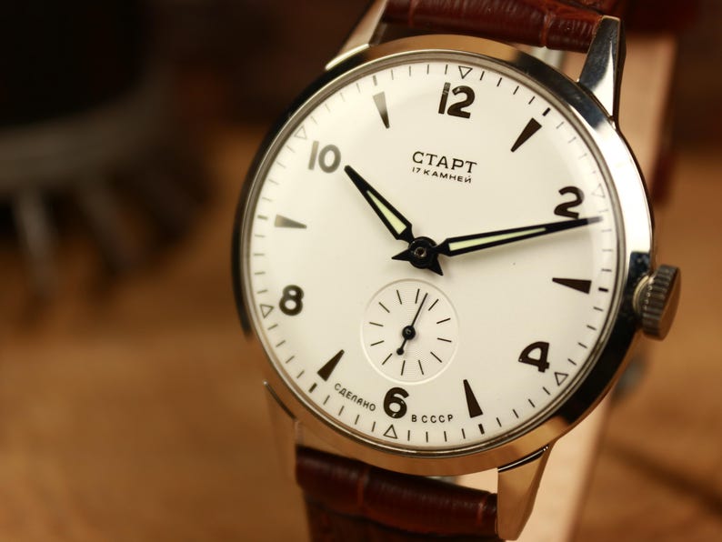 May include: A vintage wristwatch with a white face, black numerals, and black hands. The watch features a small seconds dial and a brown leather strap. The watch face displays the Cyrillic text "CTAPT" and "17 KAMHEЙ".