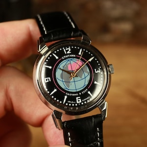 Könnte beinhalten: Eine Vintage-Armbanduhr mit schwarzem Zifferblatt und schwarzem Lederarmband. Das Zifferblatt zeigt ein Globusdesign mit dem Wort "Sputnik" und kyrillischem Text. Die Uhr hat silberfarbene Zeiger und ein silberfarbenes Gehäuse.