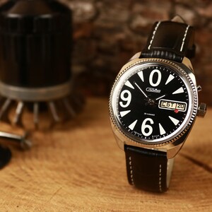 Peut inclure: Une montre-bracelet analogique noire avec des chiffres et des aiguilles blancs, et un bracelet en cuir noir. Le cadran affiche le jour et la date. La montre est posée sur un fond en bois, avec d'autres objets en arrière-plan.