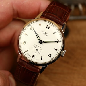 May include: A vintage wristwatch with a white face, black hands, and a brown leather strap. The watch face features the text "СТАРТ" and Arabic numerals. The watch has a small seconds dial and a stainless steel case.