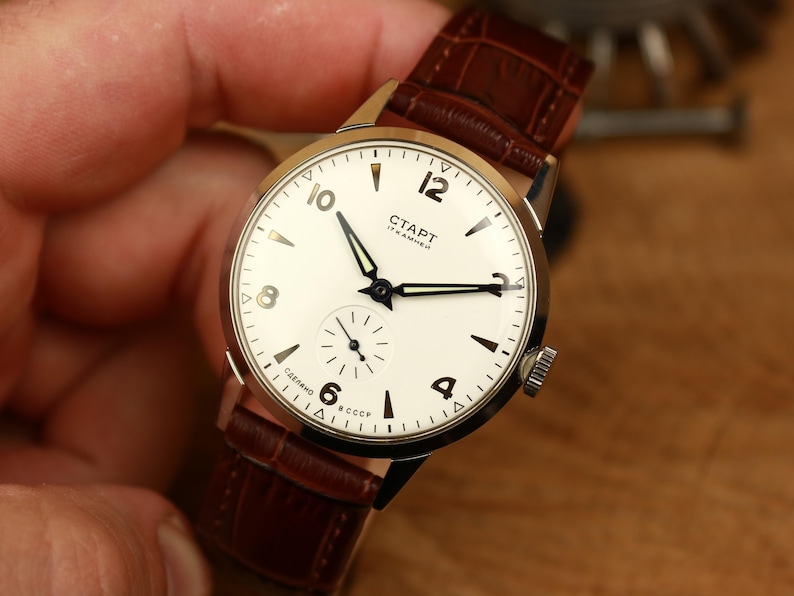 May include: A vintage wristwatch with a white face, black hands, and a brown leather strap. The watch face features the text "СТАРТ" and Arabic numerals. The watch has a small seconds dial and a stainless steel case.