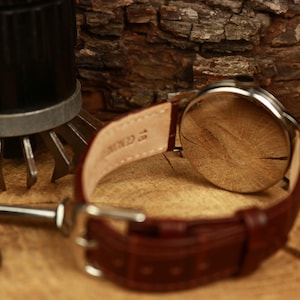 May include: A wristwatch with a brown leather strap. The watch has a silver case and a light-coloured face. The strap is fastened with a silver buckle. The watch is on a wooden surface. The strap is marked "Genuine".