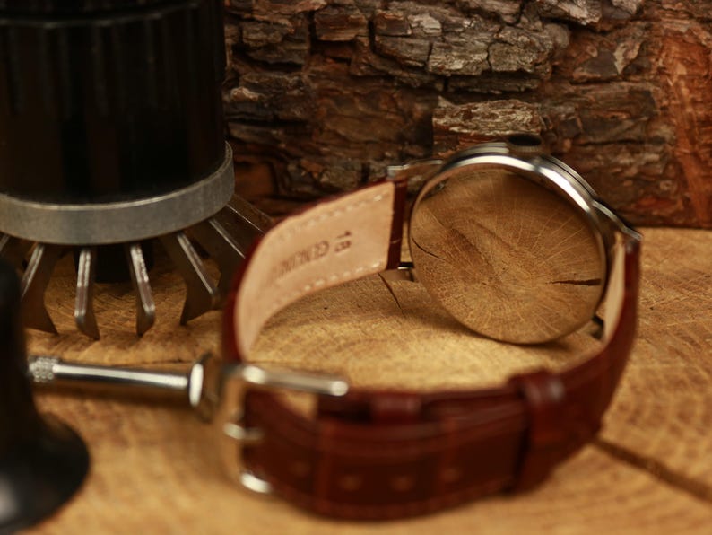 May include: A wristwatch with a brown leather strap. The watch has a silver case and a light-coloured face. The strap is fastened with a silver buckle. The watch is on a wooden surface. The strap is marked "Genuine".